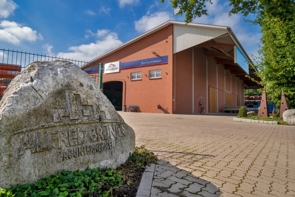 Kim Vordergrund ist ein Stein mit dem Firmenlogo zu sehen, im Hintergrund steht die Halle von Alfred Brünjes Bauunternehmen.