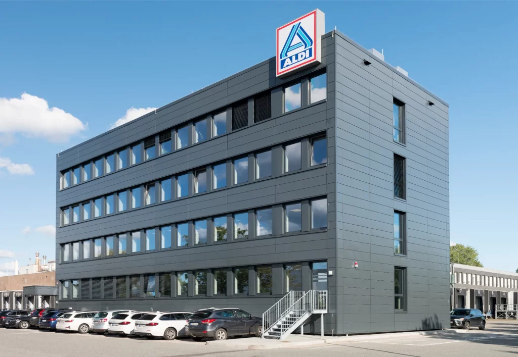 Ein ALDI Verwaltungsgebäude mit Parkplätzen. Oben rechts am Gebäude ist das ALDI Logo angebracht.