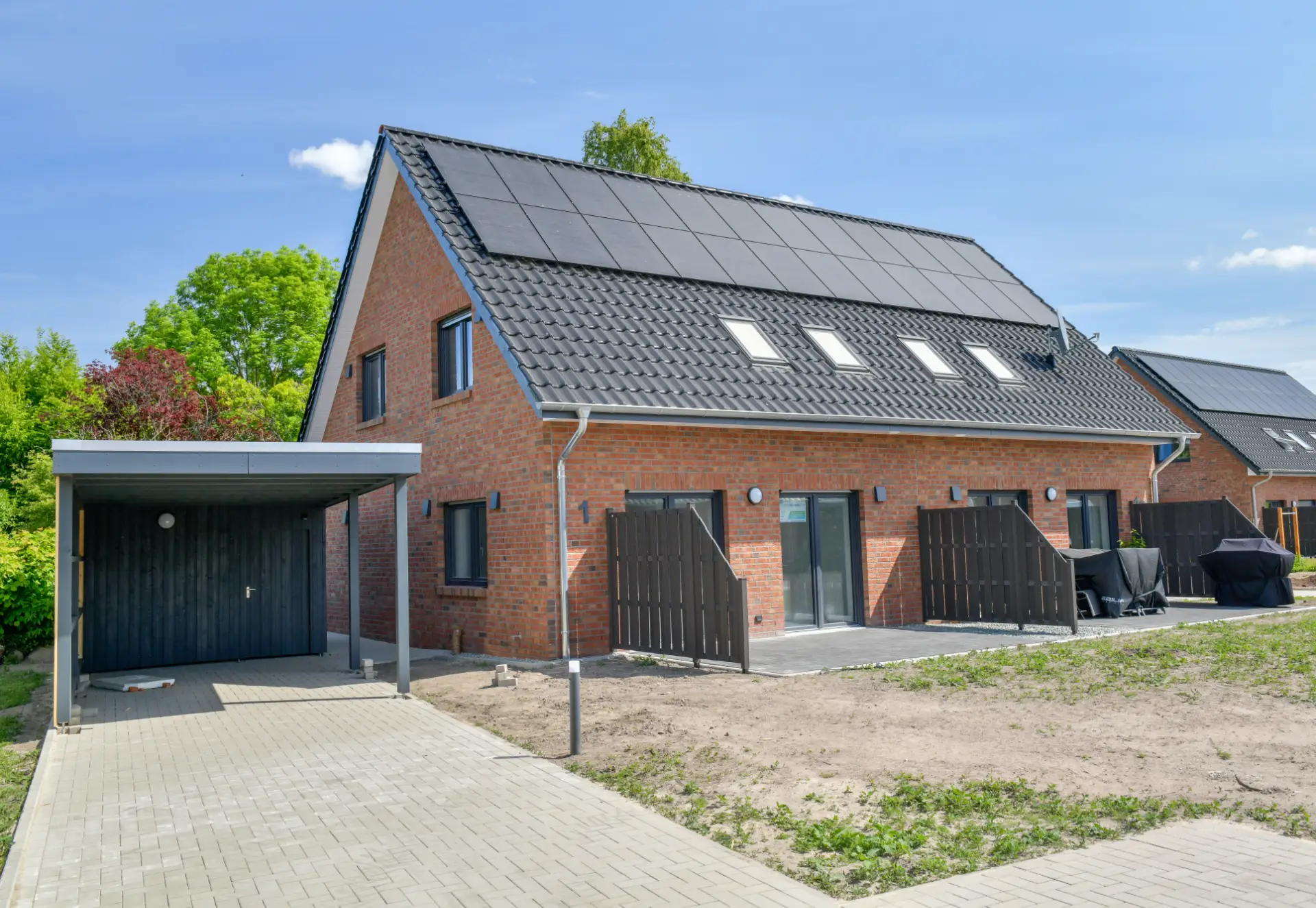 Ein Doppelhaus mit Photovoltaik auf dem Dach und Terrassen, daneben ein Parkplatz mit Überdachung.